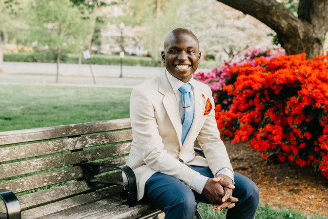 Oye Owolewa is sitting on a wooden bench in a park, dressed in a light suit with a blue tie, smiling brightly with colourful flowers in the background.