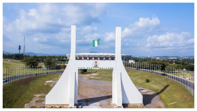 Aerial view of the Abuja City Gate with a waving Nigerian flag at the top, surrounded by lush greenery and a clear blue sky