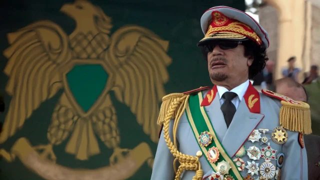 Muammar Gaddafi in a grey military uniform adorned with multiple medals and sunglasses, looking intently forward.
